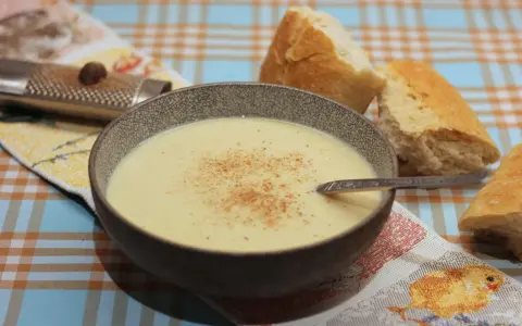 Eine Schüssel cremiger Stracciatella alla Romana, bestreut mit Zimt, neben knusprigem Brot. Ein Nussreibstein liegt auf einem bunten Tischtuch.