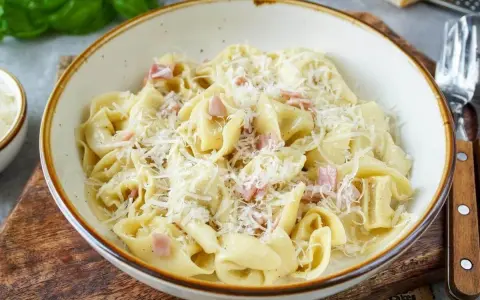 Eine Schüssel mit cremigen Tortellini alla panna, garniert mit geriebenem Parmesan und kleinen Schinkenwürfeln.