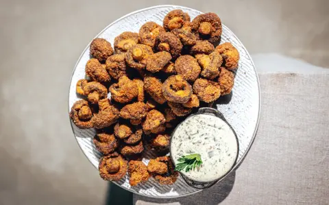 Knusprig gebackene Champignons auf einem Teller, daneben eine Schale cremiger Dip mit Kräutern. Ein Genuss!