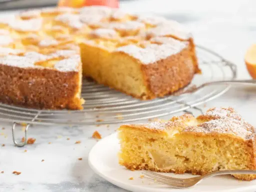 Ein saftiger glutenfreier Apfelkuchen, bestäubt mit Puderzucker, auf einem Gitterrost. Ein Stück auf einem Teller mit Gabel daneben.