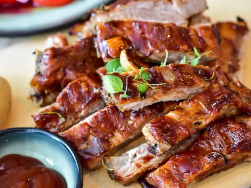 Saftige spareribs backofen, glasiert und mit Kräutern garniert, daneben eine Schale BBQ-Sauce und eine Kartoffel.