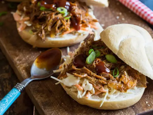 Saftiges pulled pork auf einem Brötchen mit Coleslaw und BBQ-Sauce, garniert mit Frühlingszwiebeln. Ein Löffel mit Sauce liegt daneben.