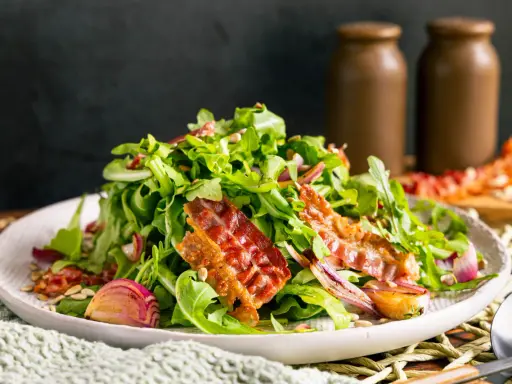 Salat mit Rucola, roten Zwiebeln und knusprigem Speck. Perfekte warme Beilagen zum Salat, die für zusätzlichen Geschmack sorgen.