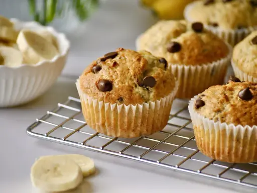 Frisch gebackene schoko bananen muffins mit Schokostückchen auf einem Gitter. Daneben eine Schüssel mit Bananenscheiben.