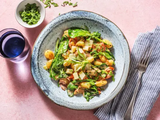Gnocchi mit Speck, Spinat und Parmesan in cremiger Sauce, serviert auf einem blauen Teller. Daneben ein Glas Wasser und eine Serviette mit Gabel.