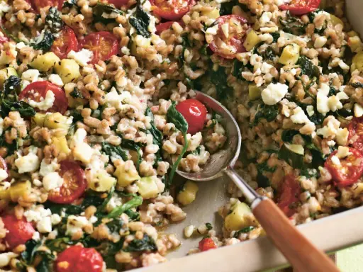 Herzhaftes Gericht mit gekochtem Dinkel, Tomaten, Zucchini und Feta. Ein Löffel hebt die köstliche Mischung aus der Auflaufform.