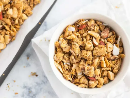 Schüssel mit selbstgemachtem, gesundem Granola, Nüssen und Kokosflocken. Daneben ein Blech mit frisch gebackenem Müsli.