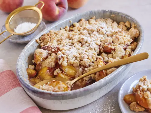 Ein goldbrauner Pfirsich Crumble mit knuspriger Streuseldecke, bestäubt mit Puderzucker. Ein Löffel steckt darin, bereit zum Genießen.