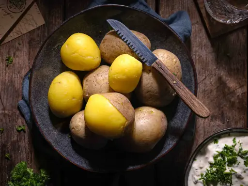 Gekochte und rohe Kartoffeln liegen in einer Schale, bereit für ein deftiges Essen. Ein Messer mit Holzgriff liegt daneben.