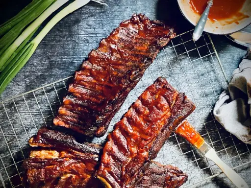 Saftige spare ribs, glasiert mit BBQ-Sauce, auf einem Grillrost. Daneben Frühlingszwiebeln und eine Schüssel Sauce.