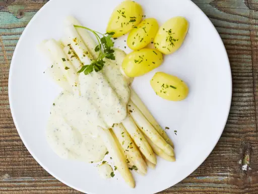 Ein Teller mit weißem Spargel, garniert mit Petersilie und überzogen mit cremiger Hollandaise. Dazu gibt es kleine, goldgelbe Kartoffeln.