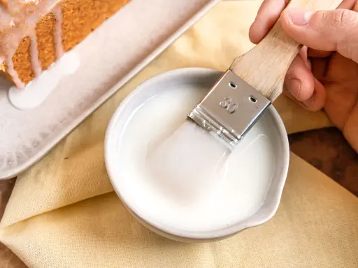 Ein Pinsel taucht in eine Schüssel mit zuckerguss, bereit, einen warmen oder kalten Kuchen zu verfeinern.