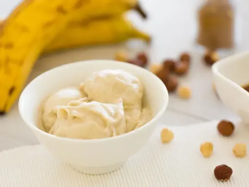 Cremiges Bananeneis, gemacht aus gefrorenen Bananen, in einer Schüssel. Daneben liegen Nüsse und ein Schälchen mit Schokoladensauce.