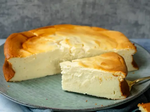 Ein saftiger Quarkkuchen ohne Boden, goldbraun gebacken, liegt auf einem Teller. Ein Stück ist bereits angeschnitten.