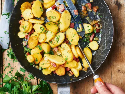 Bratkartoffeln mit mehligen Kartoffeln, Speck und Zwiebeln in einer Pfanne, garniert mit Petersilie.