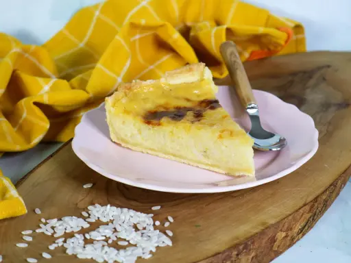 Ein Stück cremiger Milchreiskuchen auf rosa Teller, daneben roher Reis. Ein leckeres Milchreiskuchen Rezept zum Nachbacken.