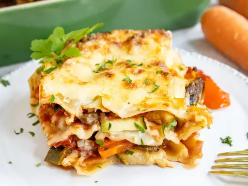 Ein Stück herzhafte vegetarische Lasagne mit geschmolzenem Käse und frischer Petersilie, daneben Karotten.