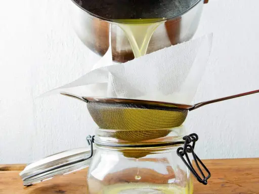 Flüssiges Ghee wird durch ein Sieb mit Küchenpapier in ein Glas gefiltert. So gelingt das Ghee selber machen.