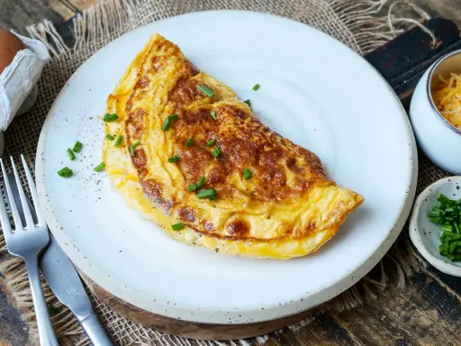 Goldbraunes Omelett auf weißem Teller, garniert mit Schnittlauch. Daneben Schüssel mit geriebenem Käse und gehacktem Schnittlauch. Perfekt für ein schnelles Omelette Rezept.