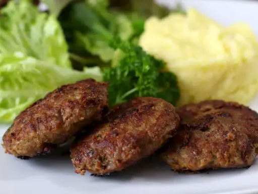 Drei goldbraune Fleischpflanzerl liegen auf einem Teller neben Kartoffelbrei und Salat. Ein leckeres Fleischpflanzerl Rezept zum Nachkochen.