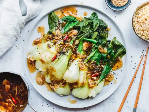 Ein Teller mit gebratenem Pak Choi, garniert mit Sesam und Chili, serviert mit Reis und einer Sauce.