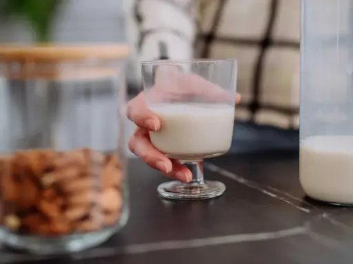 Glas Mandelmilch in Hand. Daneben ein Glas mit Mandeln und eine Flasche Milch. Man fragt sich, kann Mandelmilch schlecht werden?