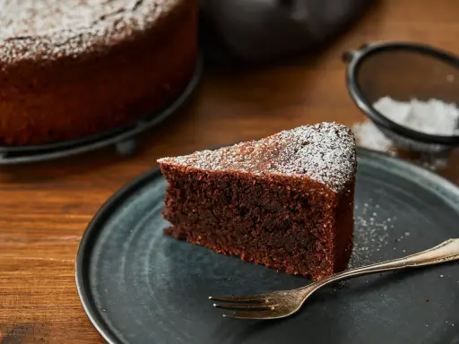 Ein Stück saftiger Schokoladenkuchen ohne Mehl, bestäubt mit Puderzucker, liegt auf einem dunklen Teller. Daneben eine Gabel und ein Sieb mit Puderzucker.