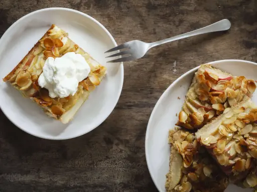 Zwei Teller mit saftigem Apfelkuchen vom Blech, bestreut mit Mandelsplittern. Eines der Stücke ist mit einer Sahnehaube garniert.