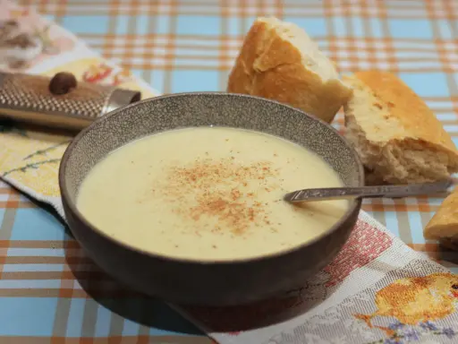 Eine Schüssel cremiger Stracciatella alla Romana, bestreut mit Zimt, neben knusprigem Brot. Ein Nussreibstein liegt auf einem bunten Tischtuch.