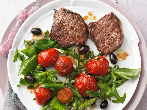 Zwei saftige Minutensteaks im Backofen zubereitet, serviert mit gerösteten Kirschtomaten, Rucola und schwarzen Oliven.