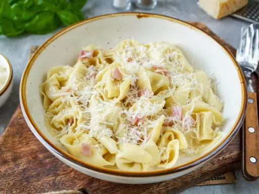 Eine Schüssel mit cremigen Tortellini alla panna, garniert mit geriebenem Parmesan und kleinen Schinkenwürfeln.