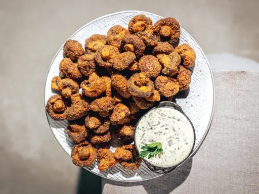 Knusprig gebackene Champignons auf einem Teller, daneben eine Schale cremiger Dip mit Kräutern. Ein Genuss!