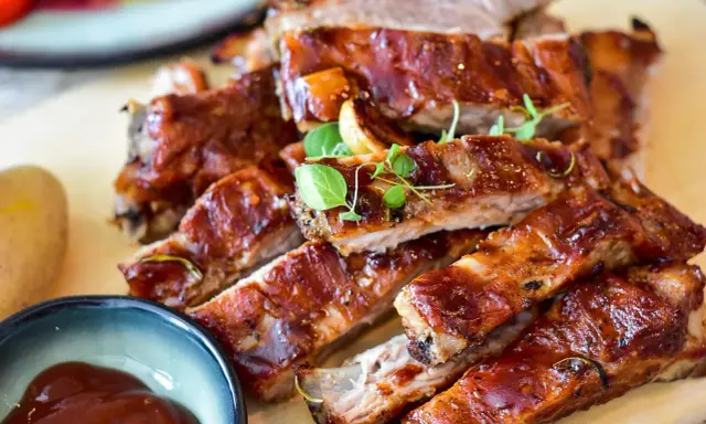 Saftige spareribs backofen, glasiert und mit Kräutern garniert, daneben eine Schale BBQ-Sauce und eine Kartoffel.