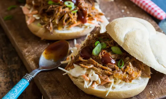 Saftiges pulled pork auf einem Brötchen mit Coleslaw und BBQ-Sauce, garniert mit Frühlingszwiebeln. Ein Löffel mit Sauce liegt daneben.