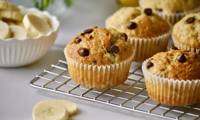 Frisch gebackene schoko bananen muffins mit Schokostückchen auf einem Gitter. Daneben eine Schüssel mit Bananenscheiben.