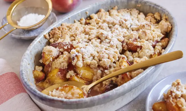 Ein goldbrauner Pfirsich Crumble mit knuspriger Streuseldecke, bestäubt mit Puderzucker. Ein Löffel steckt darin, bereit zum Genießen.