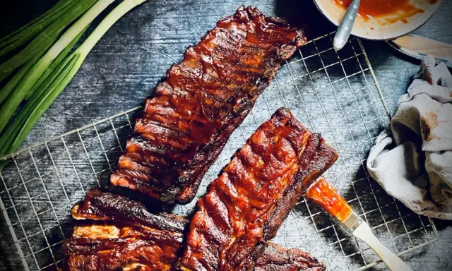 Saftige spare ribs, glasiert mit BBQ-Sauce, auf einem Grillrost. Daneben Frühlingszwiebeln und eine Schüssel Sauce.