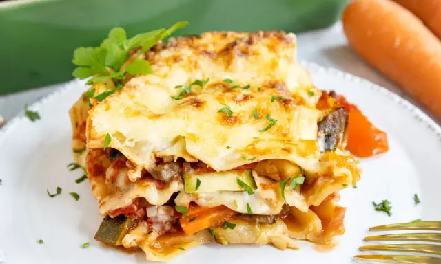 Ein Stück herzhafte vegetarische Lasagne mit geschmolzenem Käse und frischer Petersilie, daneben Karotten.