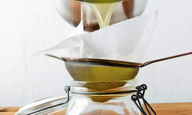 Flüssiges Ghee wird durch ein Sieb mit Küchenpapier in ein Glas gefiltert. So gelingt das Ghee selber machen.