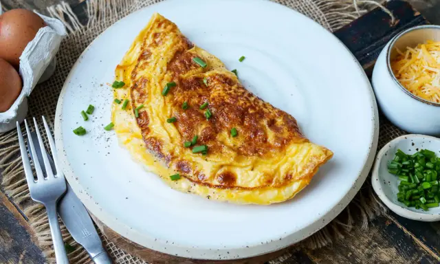 Goldbraunes Omelett auf weißem Teller, garniert mit Schnittlauch. Daneben Schüssel mit geriebenem Käse und gehacktem Schnittlauch. Perfekt für ein schnelles Omelette Rezept.