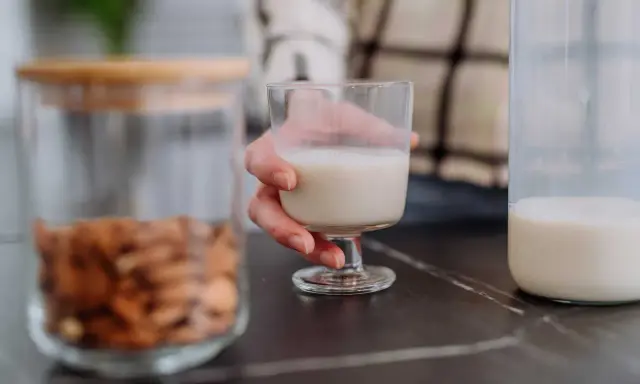 Glas Mandelmilch in Hand. Daneben ein Glas mit Mandeln und eine Flasche Milch. Man fragt sich, kann Mandelmilch schlecht werden?