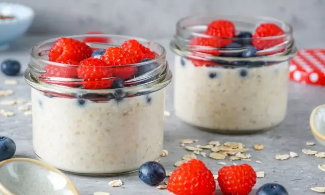 Zwei Gläser mit Overnight Oats, garniert mit frischen Himbeeren und Blaubeeren. Perfekt für ein gesundes Frühstück.