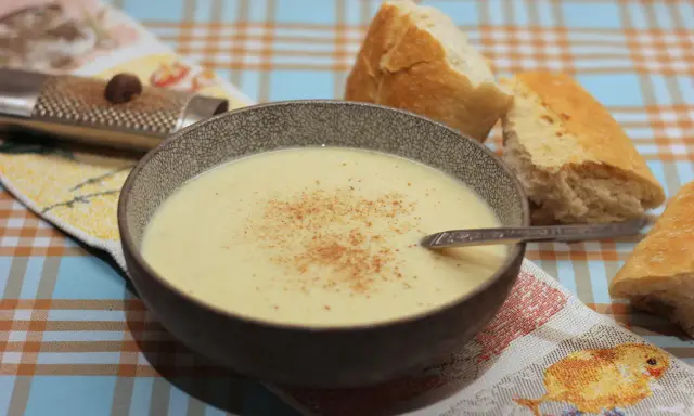Eine Schüssel cremiger Stracciatella alla Romana, bestreut mit Zimt, neben knusprigem Brot. Ein Nussreibstein liegt auf einem bunten Tischtuch.