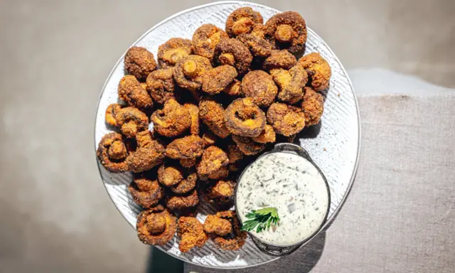 Knusprig gebackene Champignons auf einem Teller, daneben eine Schale cremiger Dip mit Kräutern. Ein Genuss!
