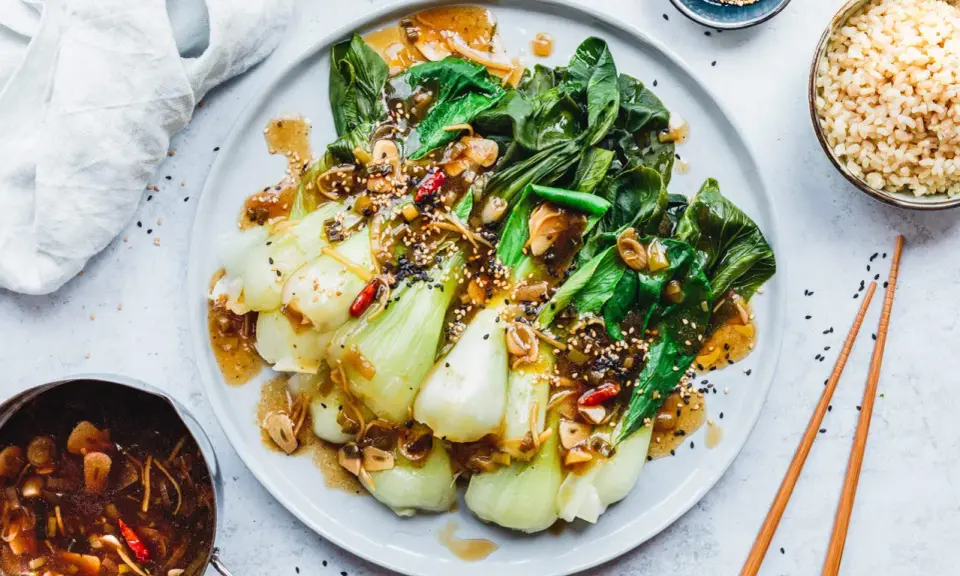 Ein Teller mit gebratenem Pak Choi, garniert mit Sesam und Chili, serviert mit Reis und einer Sauce.