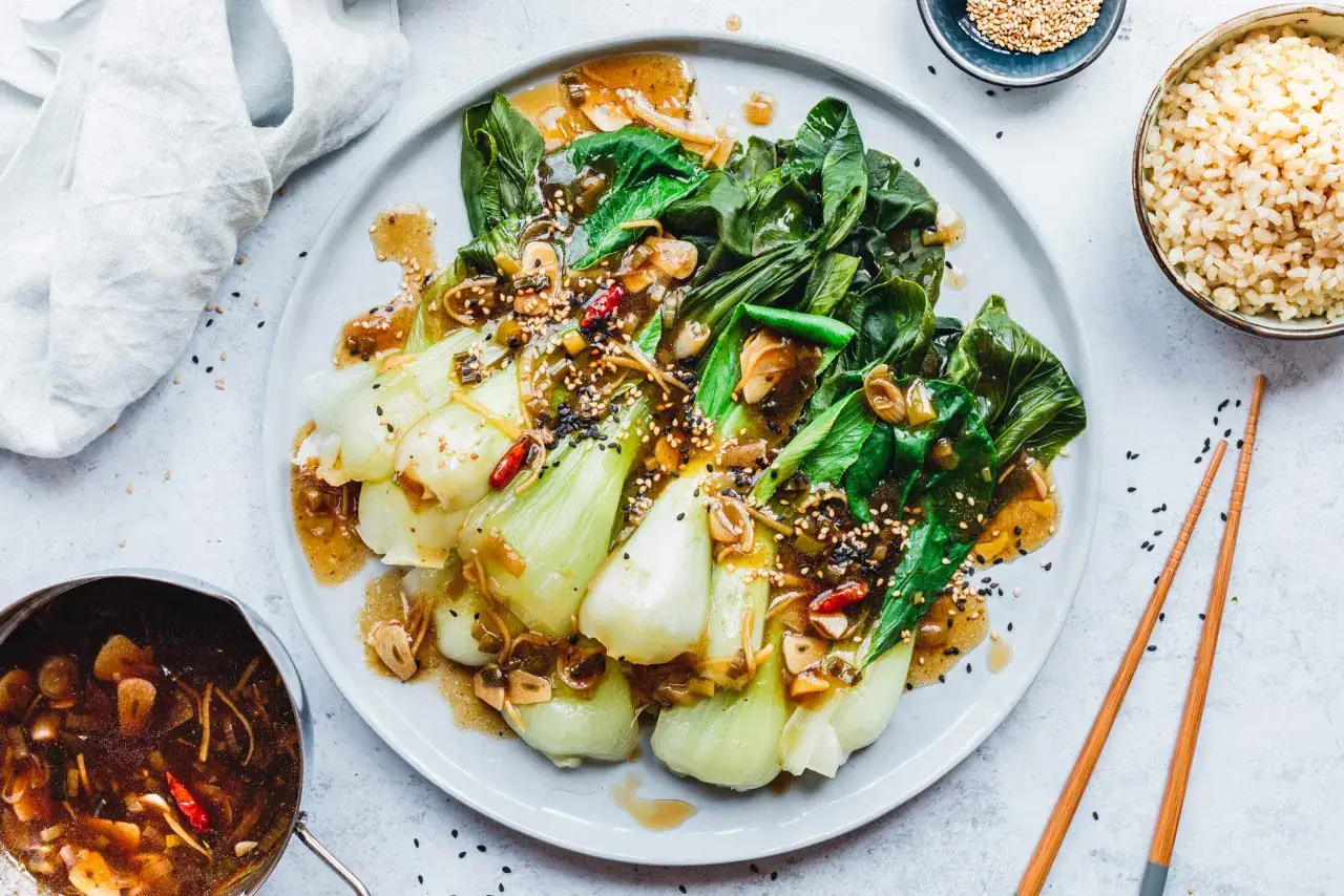 Ein Teller mit gebratenem Pak Choi, garniert mit Sesam und Chili, serviert mit Reis und einer Sauce.