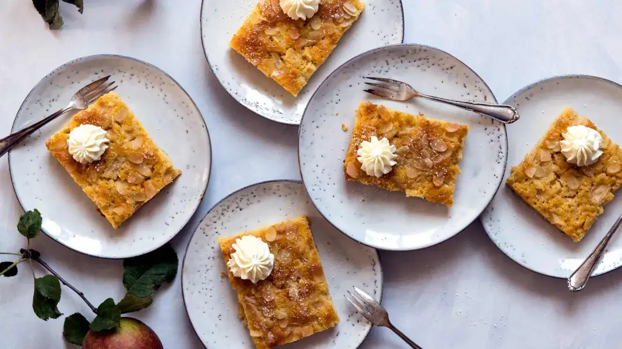Vier St&uuml;cke vom besten Apfelkuchen mit Mandelsplittern und Sahnetupfern auf Tellern, bereit zum Genie&szlig;en.