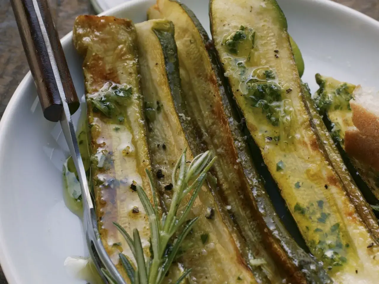 Gegrillte Zucchinischeiben mit Kr&auml;utern und &Ouml;l, perfekt zum Marinieren. Ein Rosmarinzweig liegt daneben.