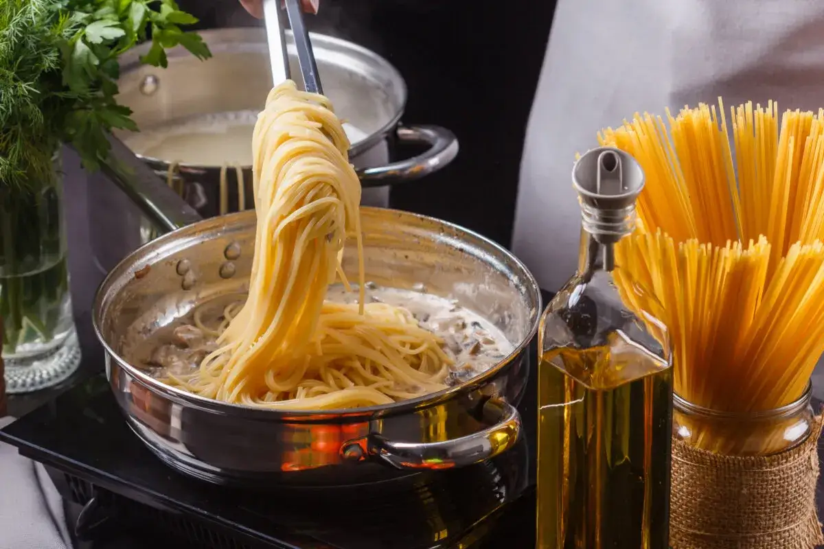 Spaghetti werden mit einer Zange aus einem Topf gehoben und in eine Pfanne mit cremiger So&szlig;e gegeben. Daneben stehen Oliven&ouml;l und trockene Spaghetti.