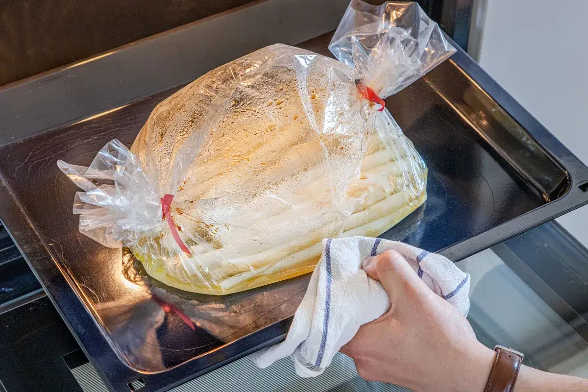 Spargel im Bratschlauch dampft auf einem Backblech. Eine Hand mit Küchentuch schiebt das Blech in den Ofen.